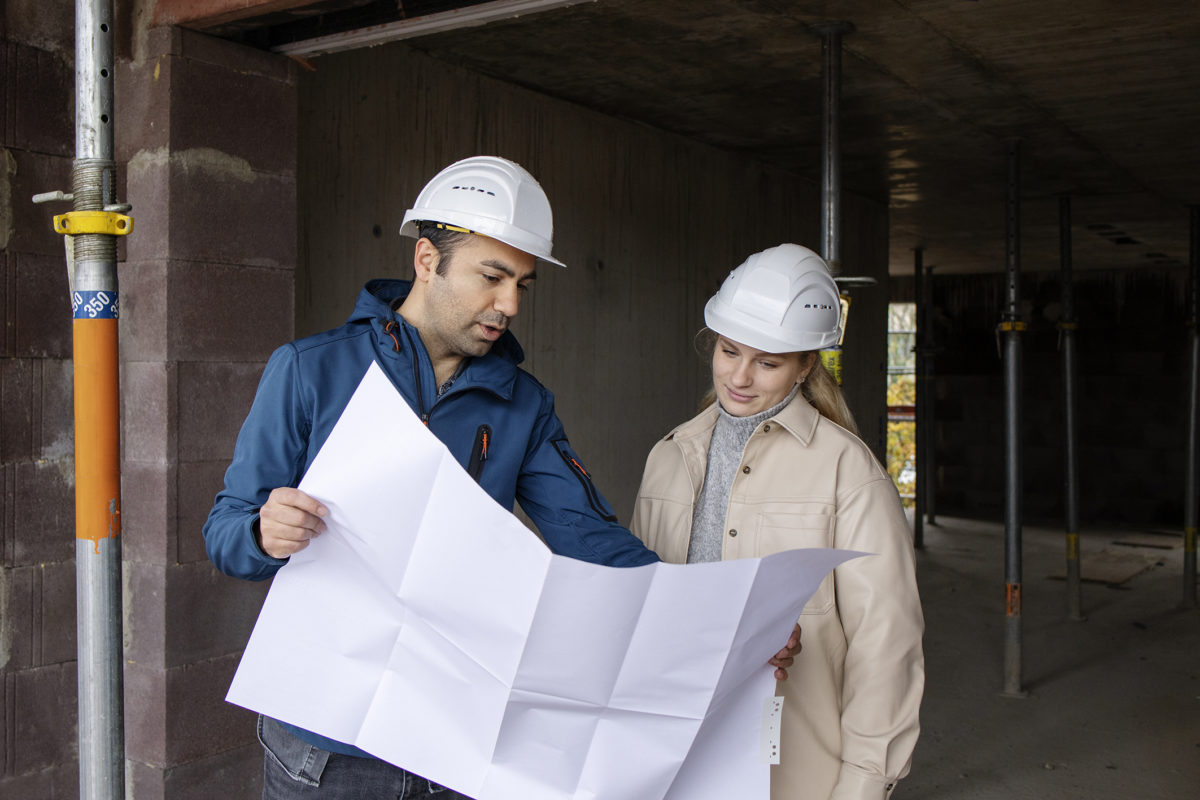 Ein Mann mit Bauhelm und eine junge Frau mit Bauhelm stehen auf einer Baustelle und besprechen einen Bauplan.
