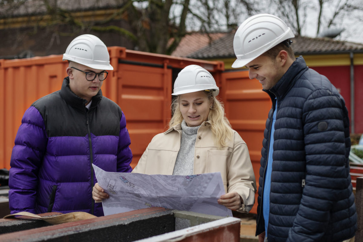 Drei junge Auszubildende der Volkswohnung stehen auf einer Baustelle und halten einen Bauplan in der Hand.