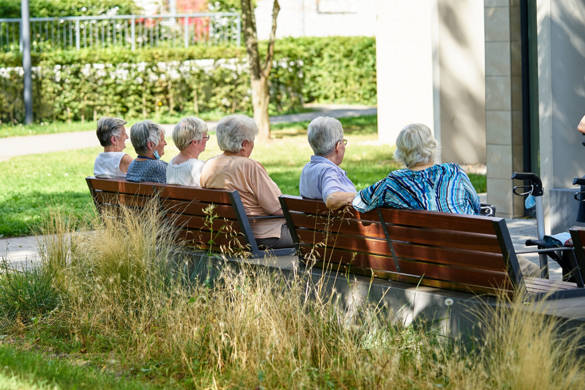 Mehrere ältere Frauen sitzen nebeneinander auf zwei Parkbänken vor einem Wohngebäude.