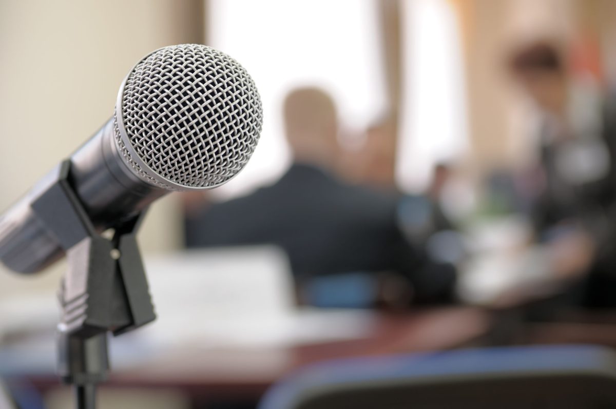 Ein Mikrofon im Vordergrund und eine Konferenz im Hintergrund unscharf.