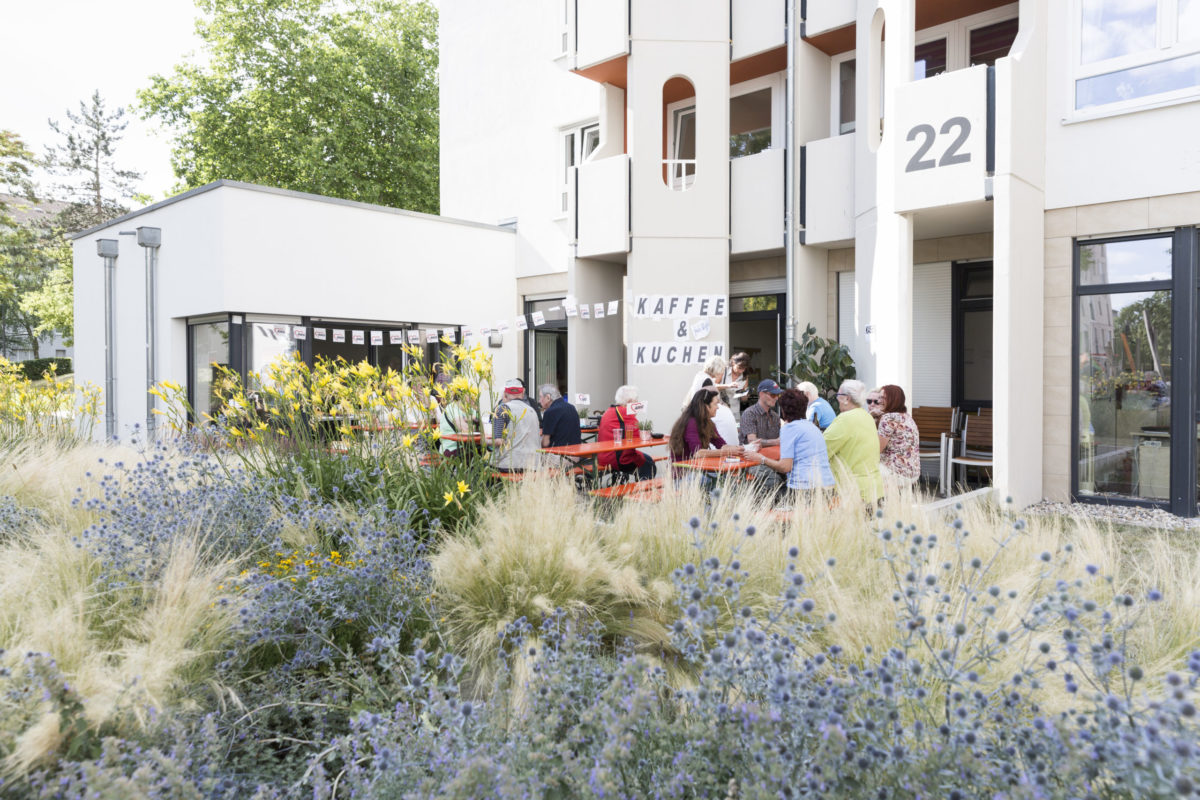 Kaffee und Kuchen genießen die Gäste bei einem Fest im AWO Wohn-Café in Rintheim. Mehrere Frauen und Männer - jung und alt - sitzen bei schönem Wetter auf Bierbänken. Im Vordergrund sieht man Gräser und blau- und gelbblühende Pflanzen.