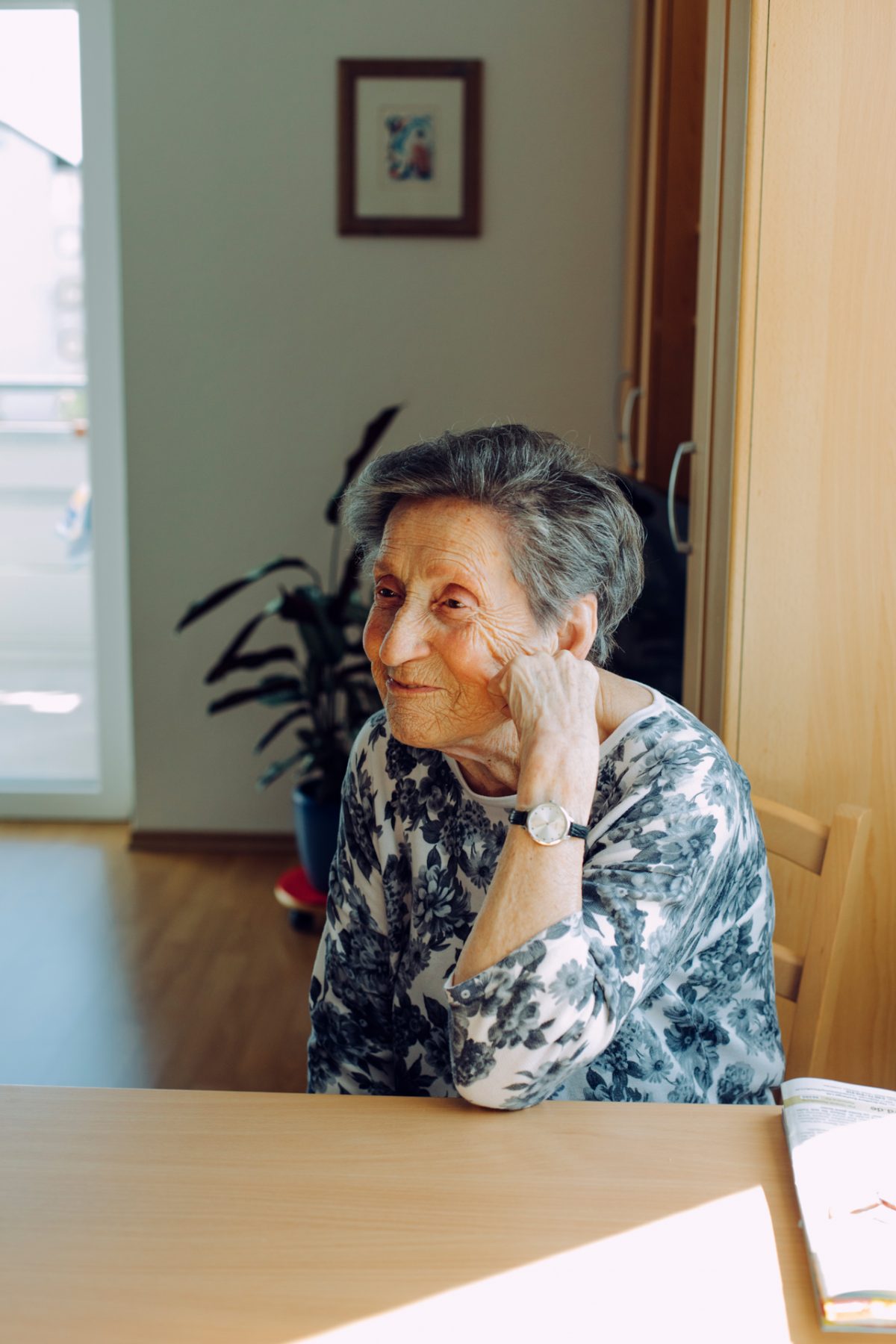 Eine ältere Dame sitzt verschmitzt an einem Tisch und stützt den Kopf auf.