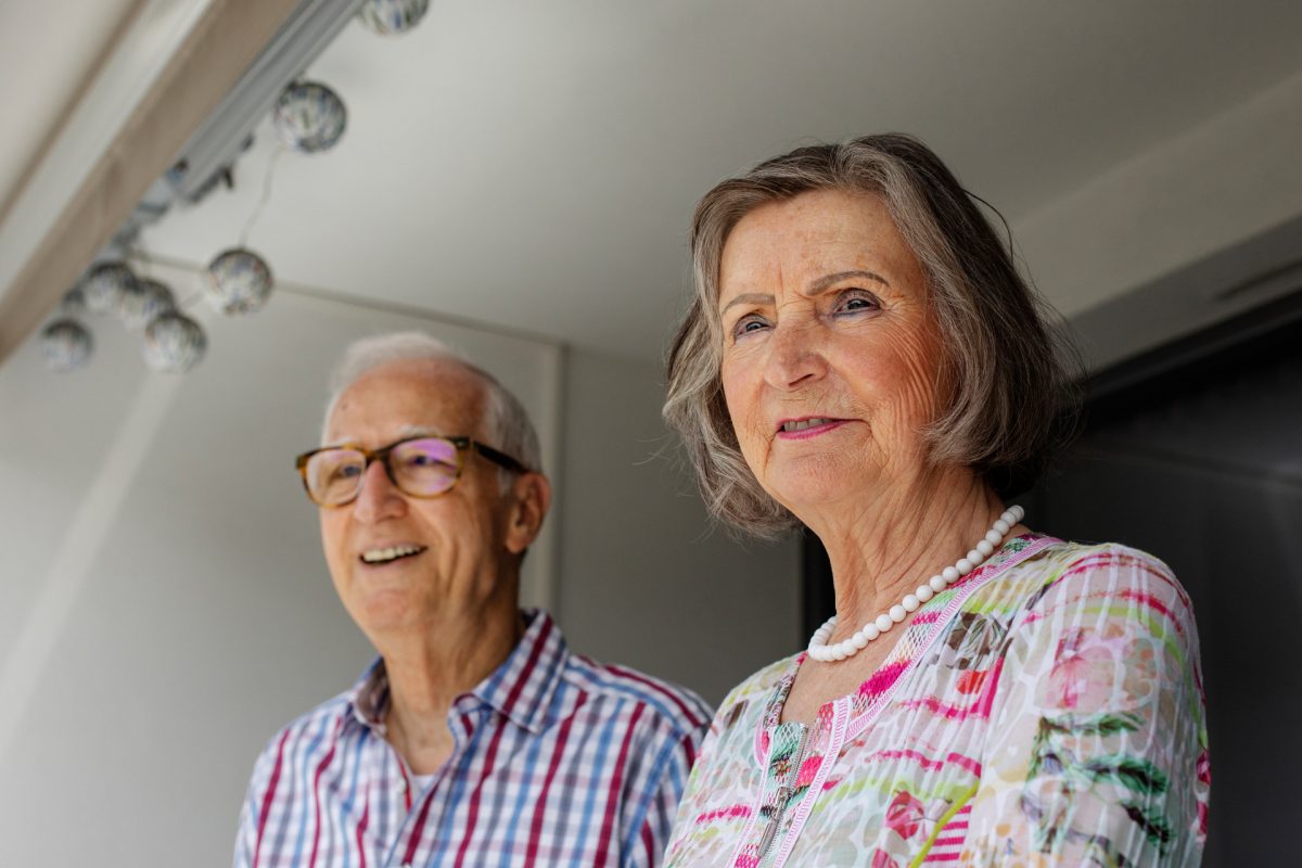 Ein älteres Ehepaar steht auf dem Balkon und blickt in die Ferne.