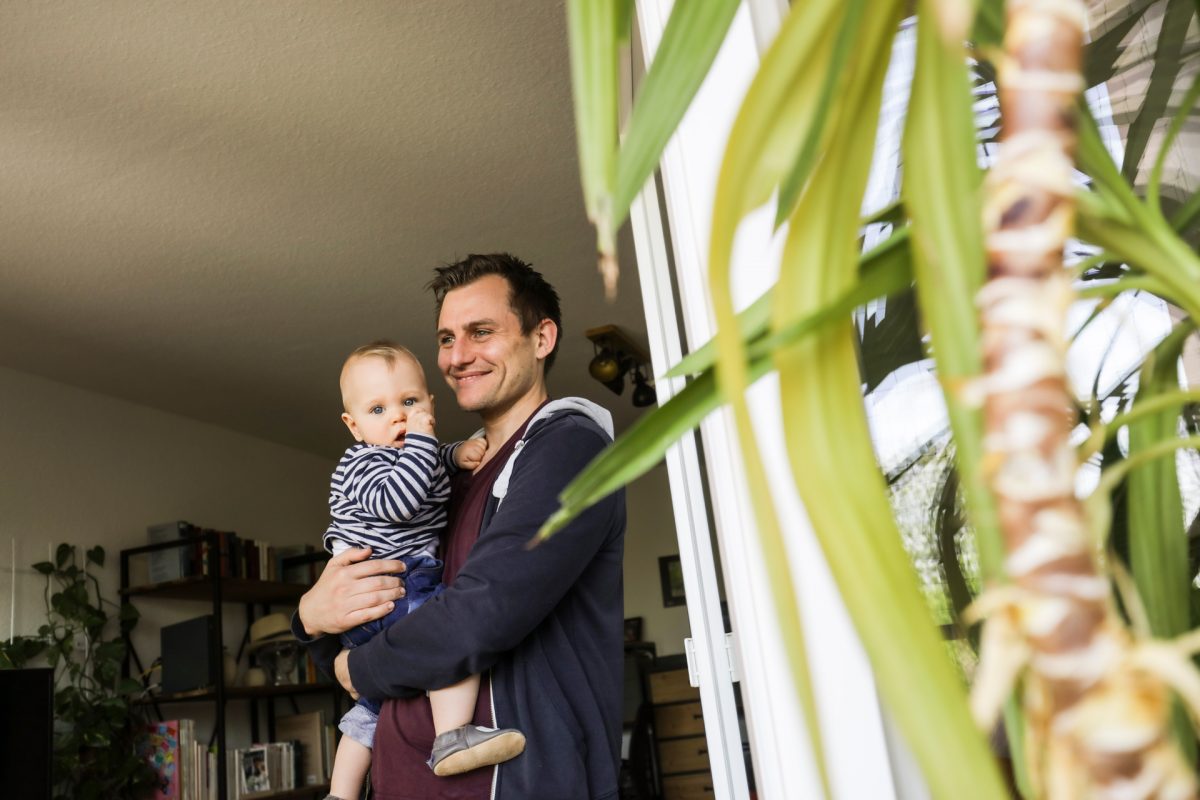 Ein junger Vater steht in der Wohnung und hält ein Kleinkind auf dem Arm.