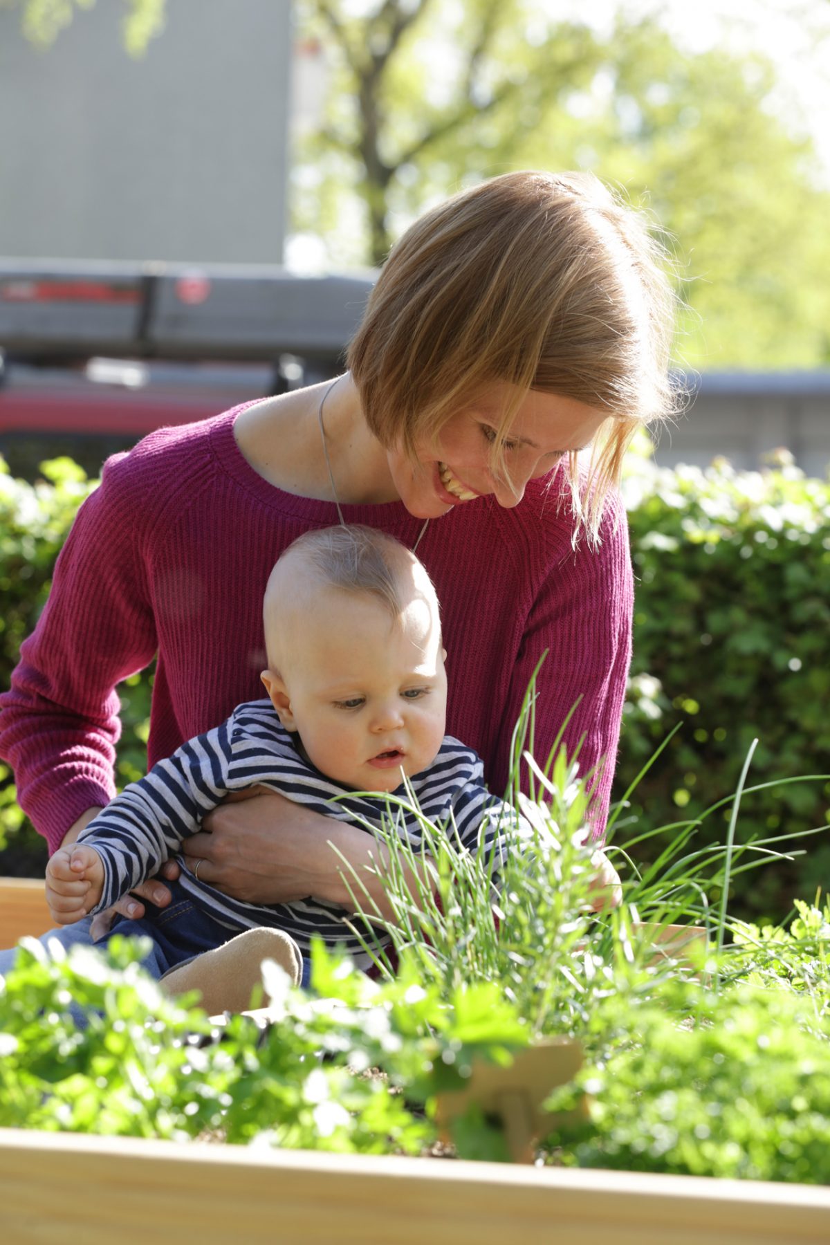 Eine junge Mutter hält ihr Kleinkind im Arm und zeigt ihm Kräuter im Hochbeet.