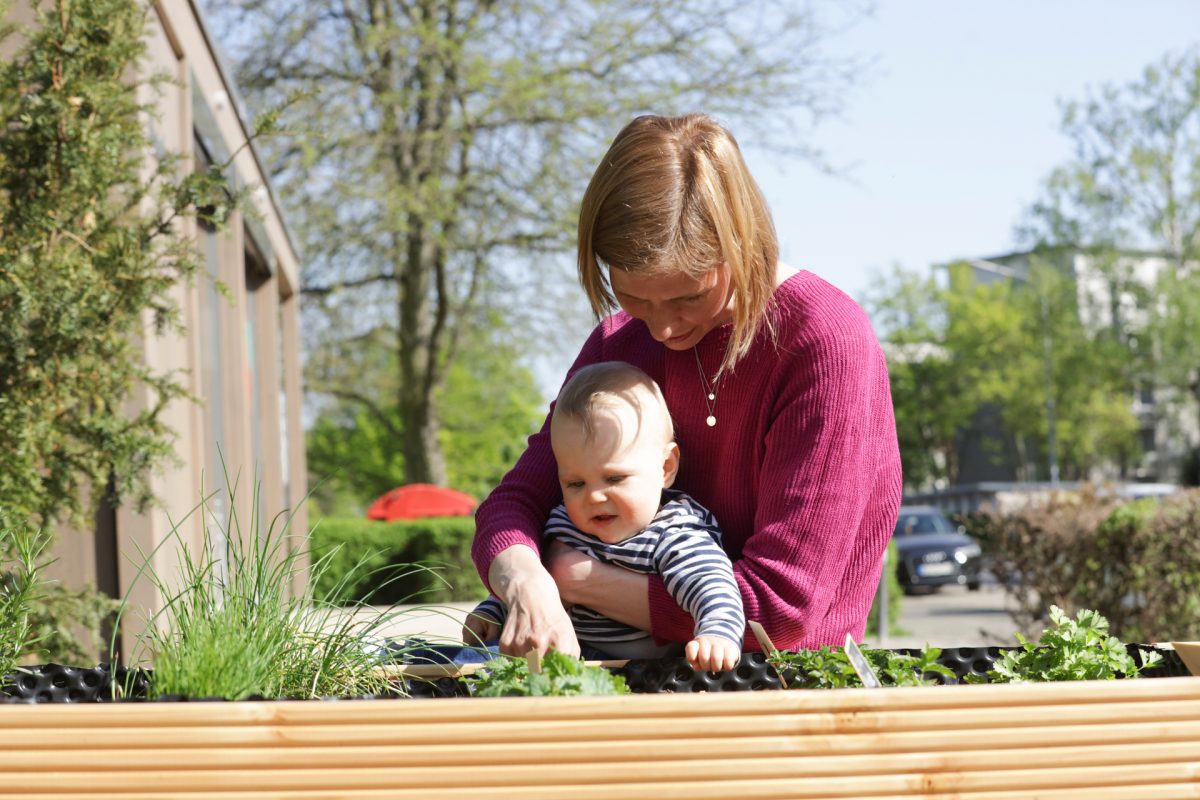 Eine junge Mutter hält ihr Kleinkind im Arm und zeigt ihm Kräuter im Hochbeet.