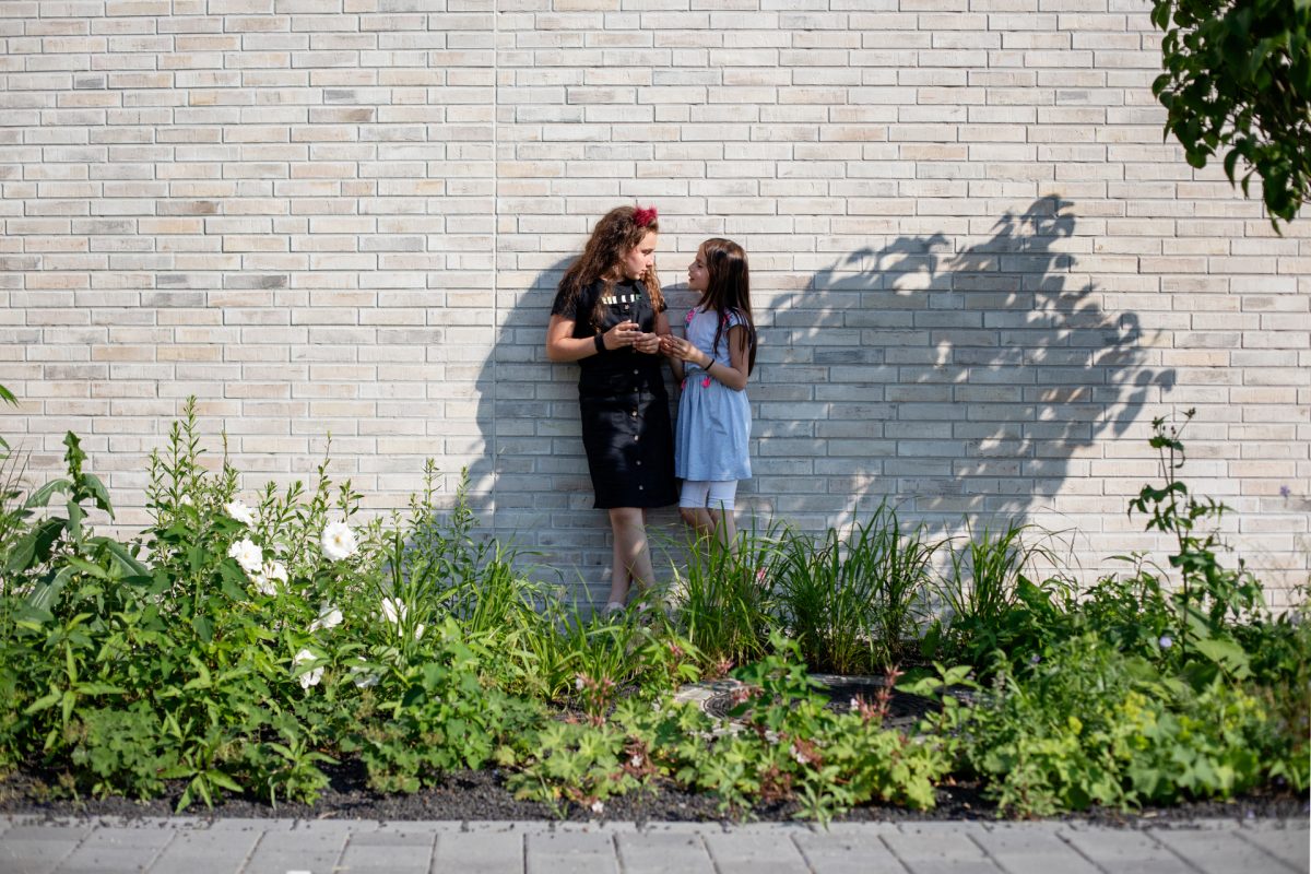 Zwei kleine Mädchen stehen an einer Mauer hinter einem bepflanzten Grünstreifen.