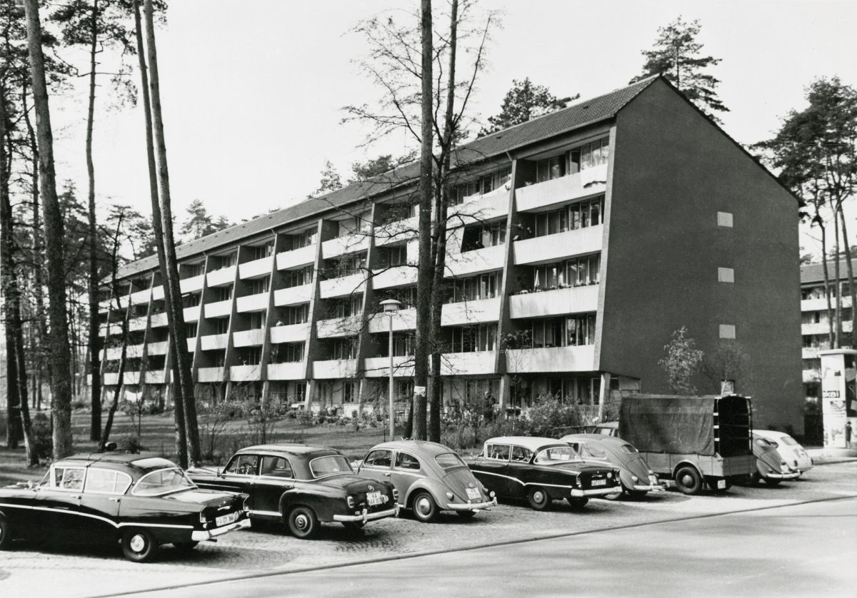 Die historische Aufnahme zeigt einen Zeilenbau in der neu entstandenen Waldstadt. Vor dem Gebäude parken Autos.