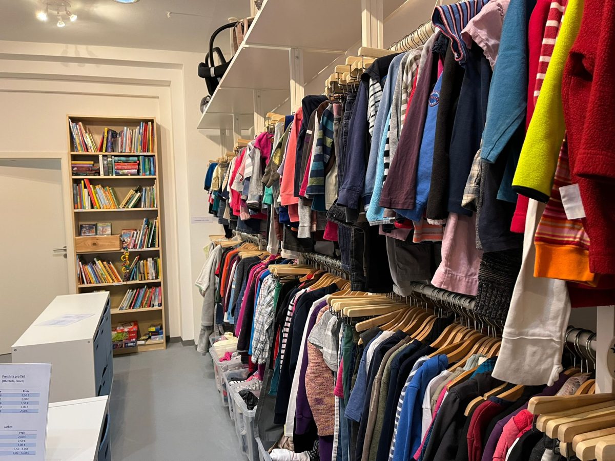 Kleiderkammer mit vielen Kleidungsstücken an Kleiderstangen, im Hintergrund ein gefüllltes Bücherregal.