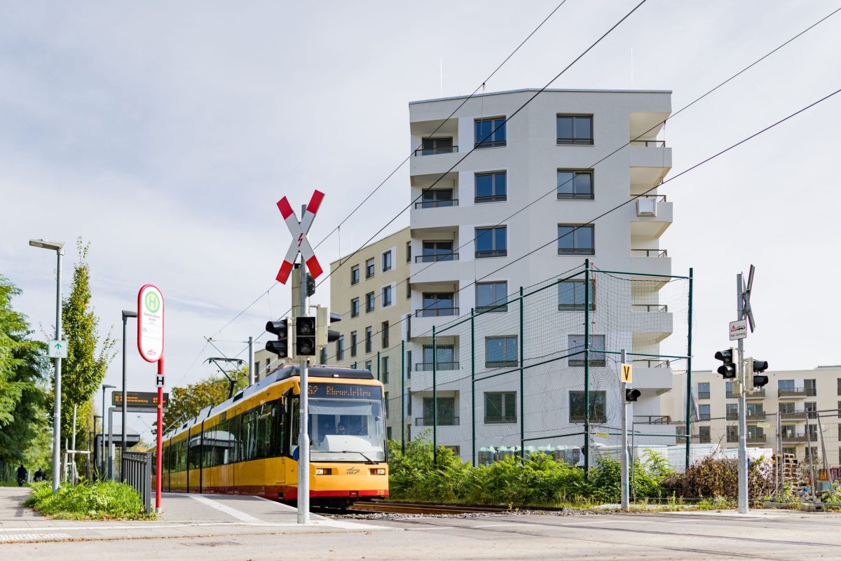 Eine Straßenbahn fährt duch die Haltestelle am August-Klingler-Areal. Im Hintergrund steht ein Gebäude des Areals.