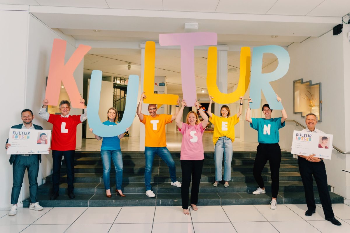 Acht Personen stehen auf einer Treppe und halten die Buchstaben K, U, L, T, U, R in die Höhe. Gefeiert wird der Neustart des Projekts Kulturlotsen.