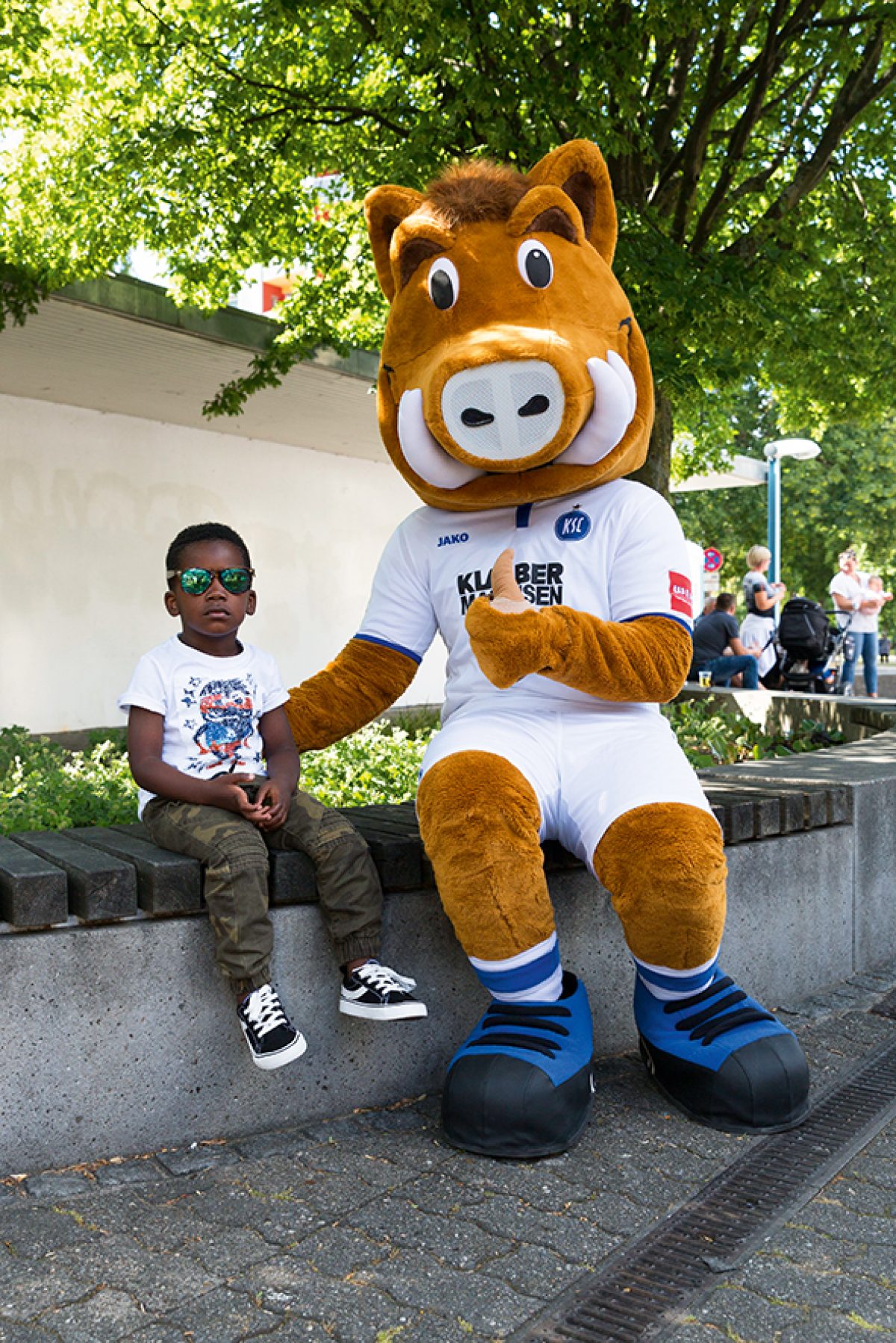 Ein kleiner Junge mit Sonnenbrille sitzt mit dem riesigen Maskottchen Wili Wildpark auf einer Mauer.