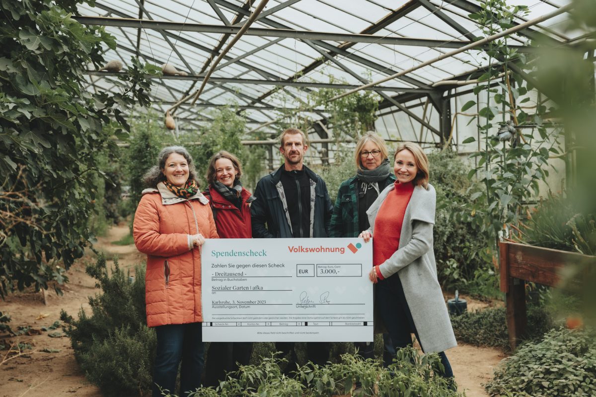 Fünf Personen stehen im Gewächshaus der Arbeitsförderungsbetriebe Karlsruhe. Der symbolische Spendenscheck wird von Pia Hesselschwerdt übergeben.