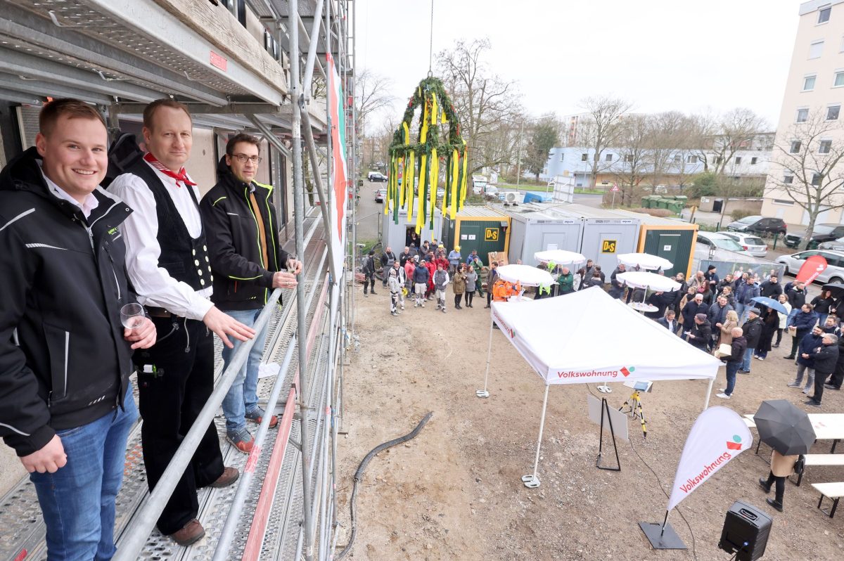 Ein Zimmermann und zwei Handwerker stehen beim Richtfest auf dem Gerüst. Der Richtkranz hängt am Kran.