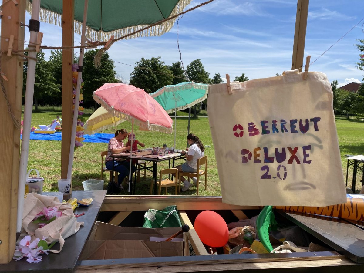 Oberreut Deluxe 2.0 steht auf einer selbst bemalten Stofftasche. Im Hintergrund sitzen Erwachsene und Kinder und bemalen weitere Taschen.