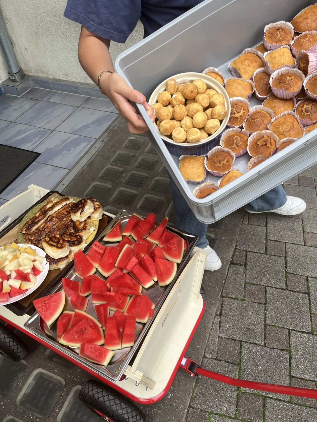 Eine Person hält eine Kiste in der Hand. Darin sind Muffins und Brötchen. Daneben gibt es auf einem Rollwagen noch ein Tablett mit Melonenstücken und anderem Fingerfood.