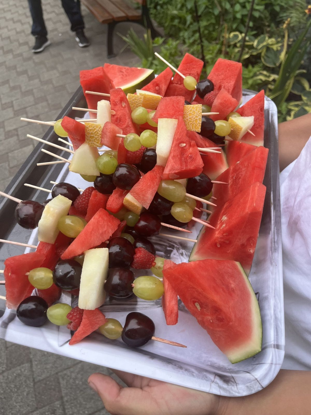 Eine Person hält ein Tablett in der Hand. Auf dem Tablett liegen Fruchtspieße.