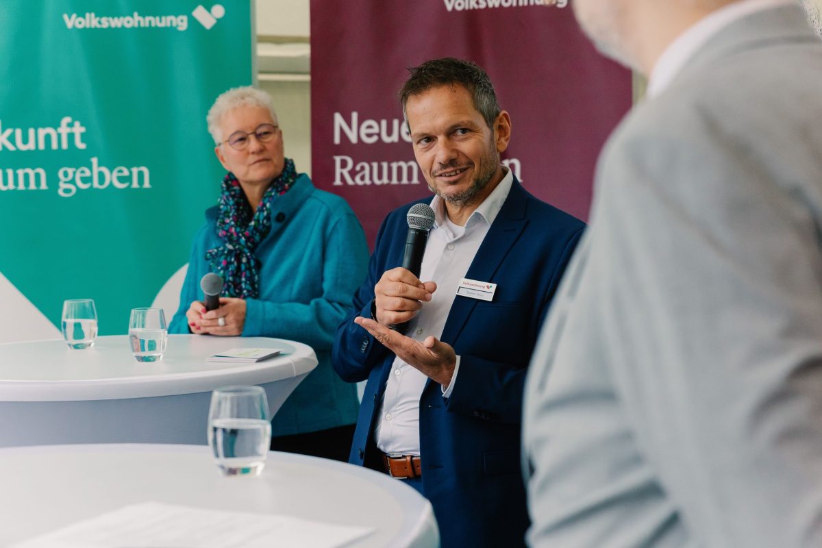 Geschäftsführer der Volkswohnung Stefan Storz während einer Podiumsdiskussion. Er spricht ins Mikrofon, hinter ihm stehen Roll-Ups der Volkswohnung.