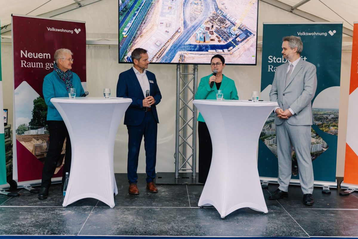 Sicht auf die Bühne bei der Fertigstellungsfeier des August-Klingler-Areals in Karlsruhe Daxlanden. Frau Annet Jura, Frau Ellen Winkler und Herr Stefan Storz sind in einer Posiumsdiskussion die vom Moderator Jan Hauser moderiert wird. Zwei Stehtische stehen im Vordergrund, im Hintergrund Rollups und ein Bildschirm.
