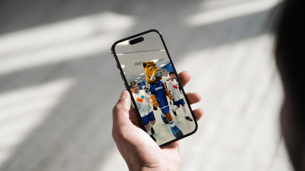 Mock up eines Smartshones. Darauf sieht man das Maskottchen vom KSC mit Einlaufkindern, die T-Shirts von der Volkswohnung tragen.