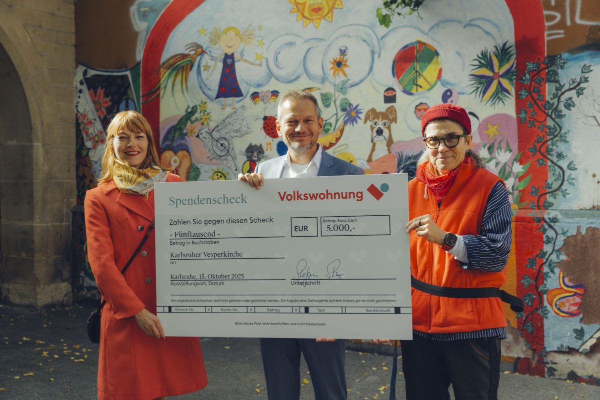 Drei Personen stehen vor einem bunten Wand-Tattoo und halten einen großen Spendenscheck in den Händen.