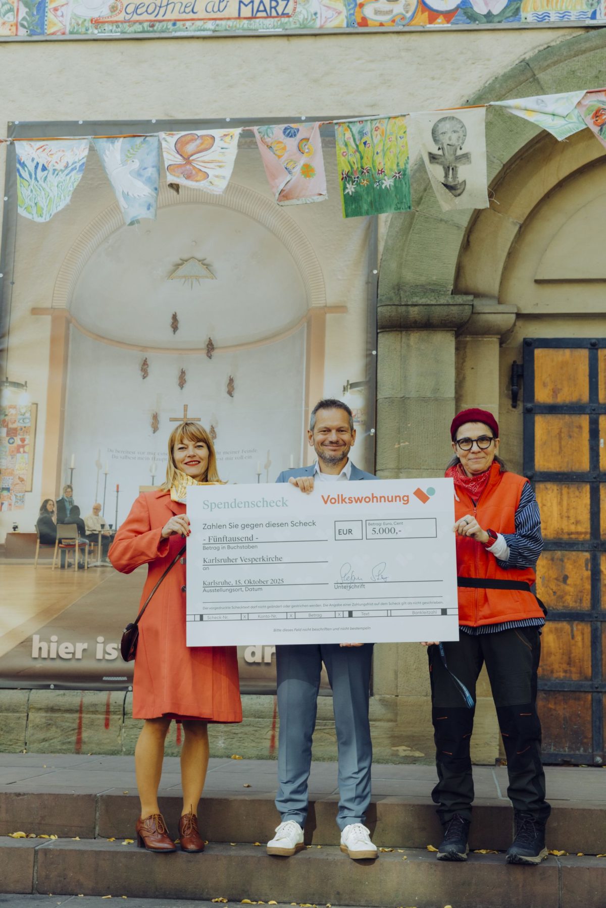 Drei Personen stehen vor auf einer Kirchentreppe und halten einen großen Spendenscheck in den Händen.