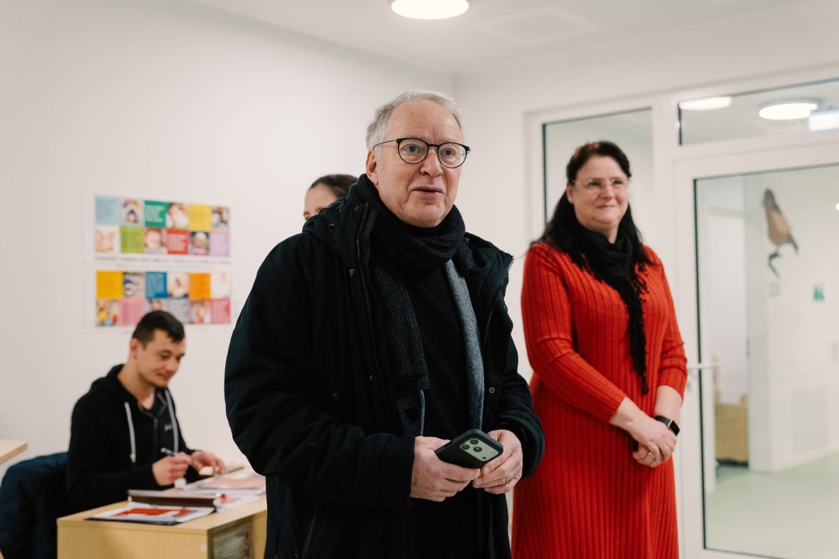 Zwei Personen stehen im Vordergrund und berichten, im Hintergrund sitzt ein Mann am Tisch.