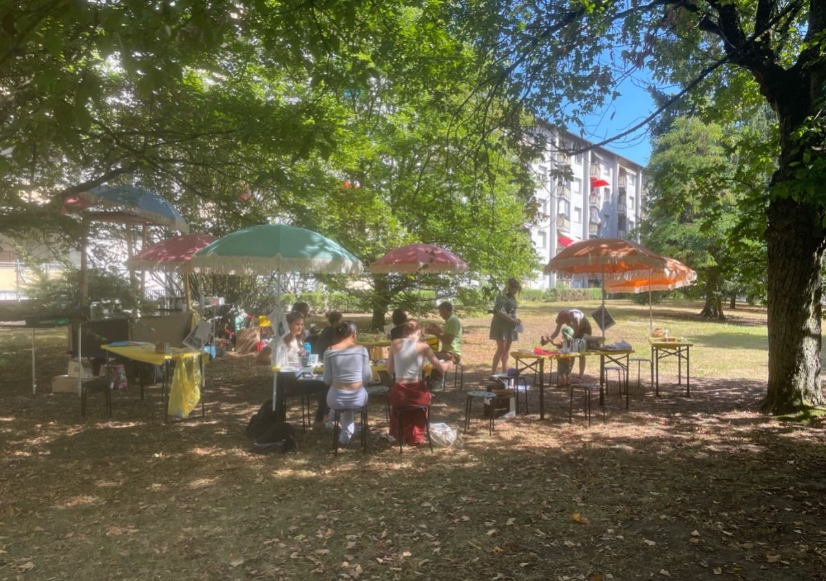 Mehrere Menschen haben auf Hocker, welche auf einer Wiese stehen Platz genommen. Sie sitzen an Festzelttischen und basteln. Sonnenschirme sind auch auf der Wiese verteilt.