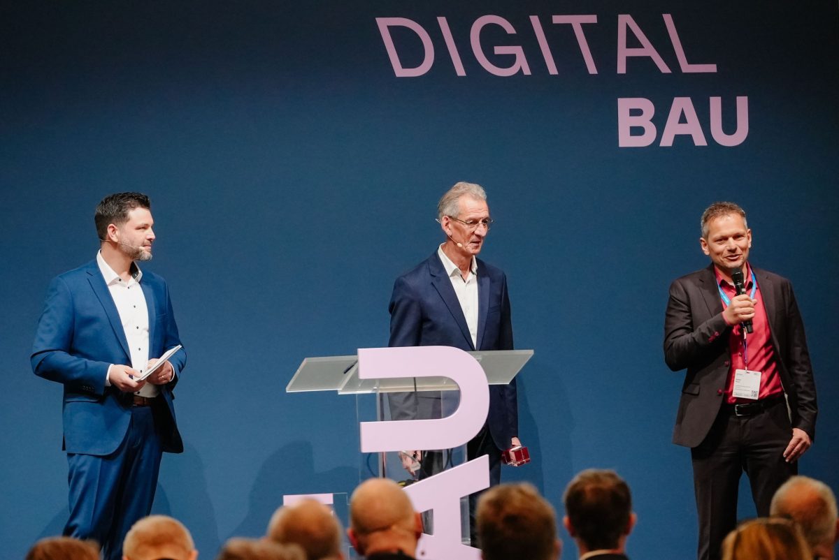 Stefan Storz, GF der Volkswohnung, bei der Preisverleihung auf der Bühne. Zu sehen sind auch der Moderator und ein Jurymitglied des Deutschen Baupreises.