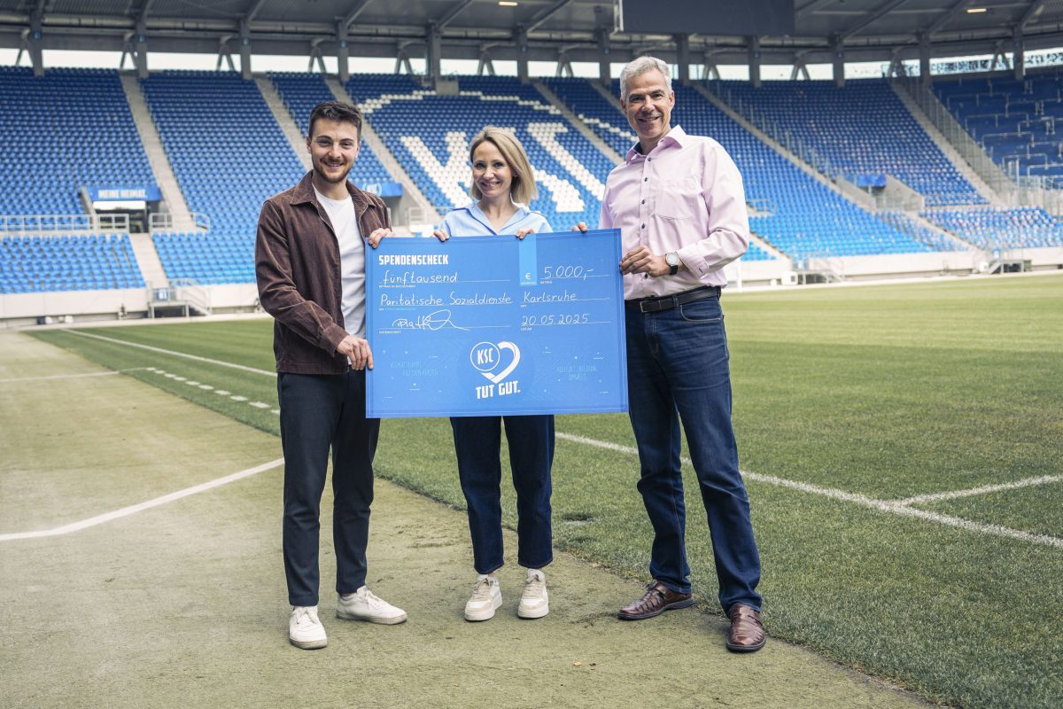 Drei Personen stehen in einem Stadion und halten einen übergroßen Spendenscheck in der Hand.