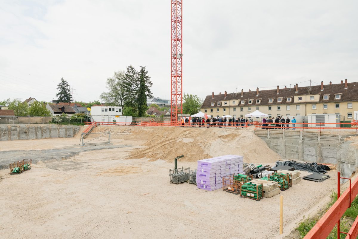 Eine große Baugrube, im Hintergrund eine Festgesellschaft anlässlich der Grundsteinlegung.