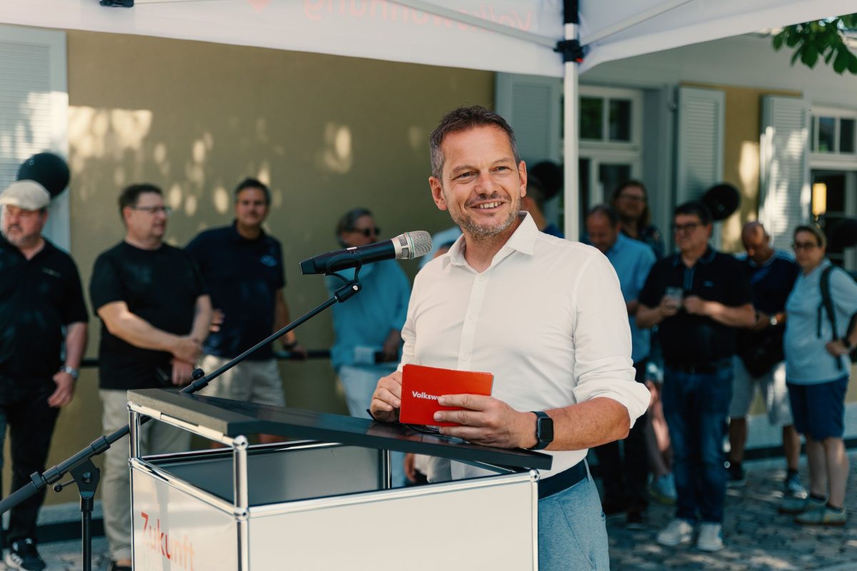 Stefan Storz, Geschäftsführer der Volkswohnung steht hinter einem Rednerpult und hält rote Karteikarten in der Hand. Im Hintergrund stehen Zuhörer:innen und hören seiner Rede zur Eröffnung des Samocca-Augustine, dem inklusiven Quartierscafé im August-Klinger-Areal, zu.
