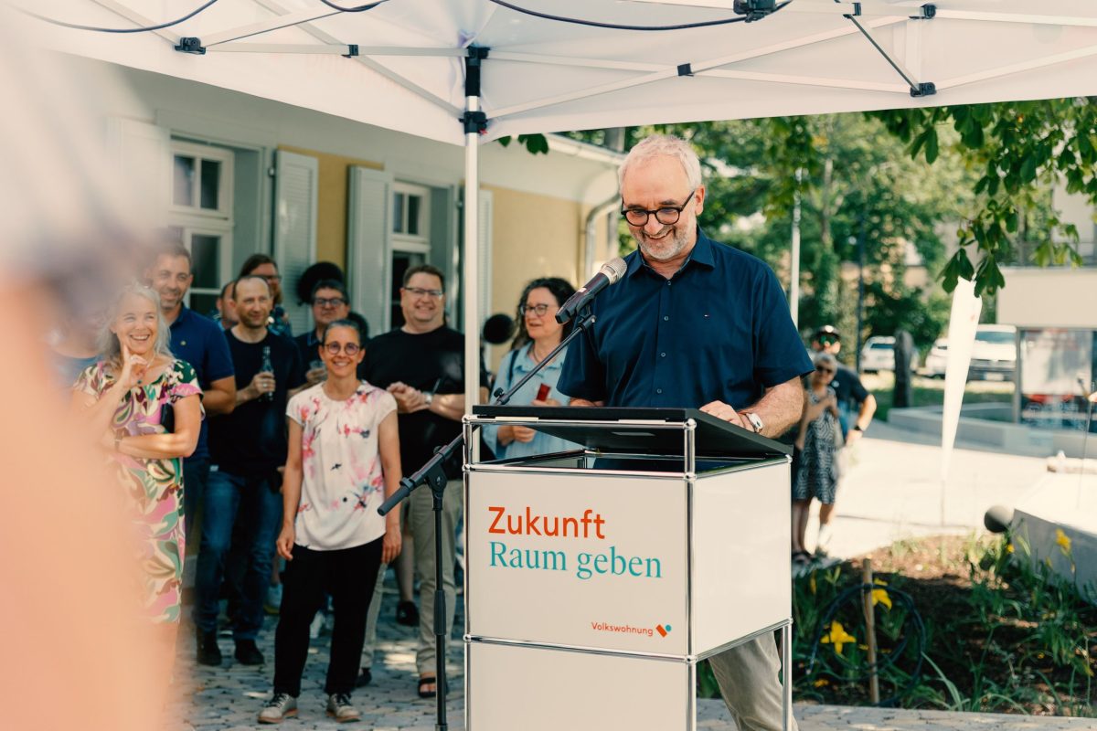 Michael Auen, Vorstand der Lebenshilfe Karlsruhe steht hinter einem Rednerpult und hält seine Rede zur Eröffnung des Samocca-Augustine. Im Hintergrund stehen Zuhörer:innen