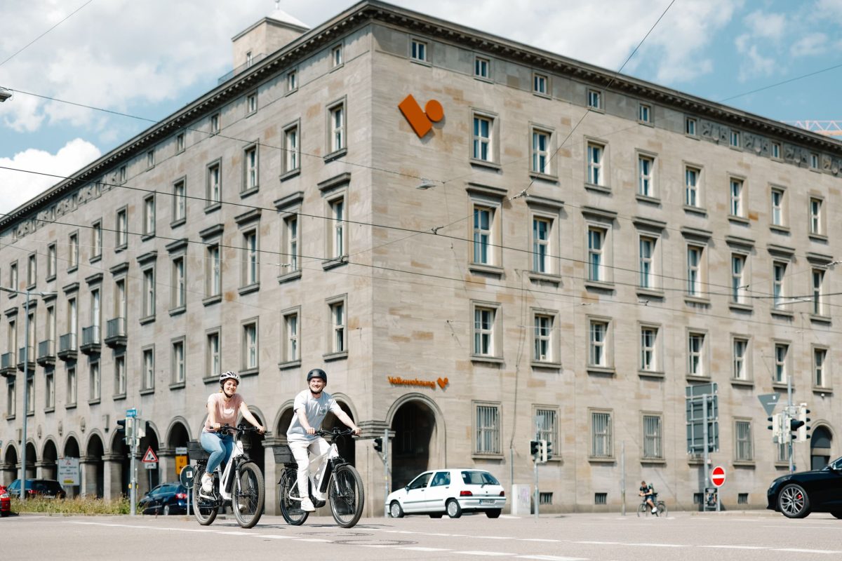 Zwei Personen fahren mit dem Rad auf der Straße. Im Hintergrund ist die Zentral der Volkswohnung.
