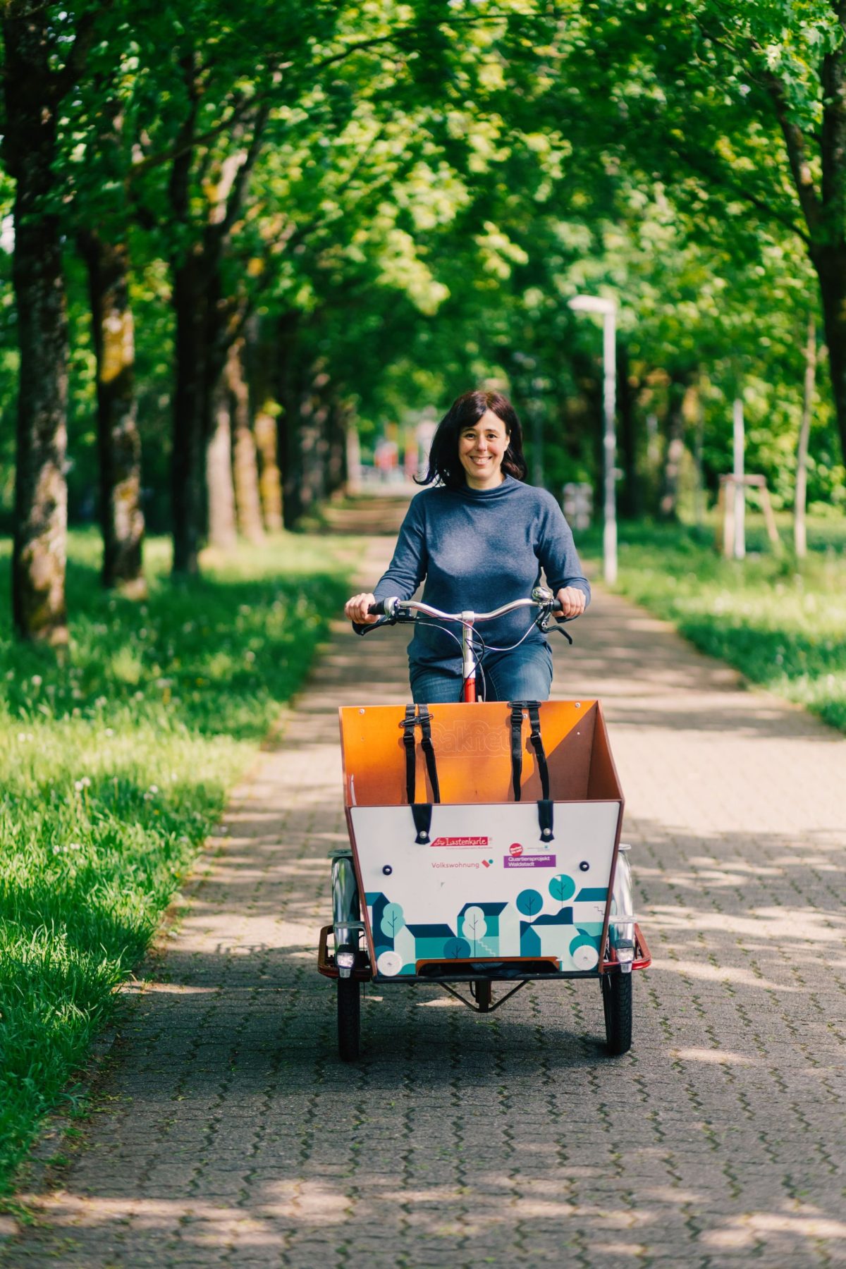An einem sonnigen Tag fährt eine Frau mit einem Lastenrad einen von Bäumen gesäumten Weg entlang und lächelt dabei in die Pedale.