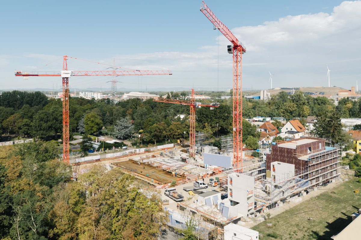 Baustelle im Buschwiesenweg mit drei hohen Kränen, mehreren Gebäuden im Bau und Gerüsten, umgeben von Grünflächen und nahegelegenen Häusern unter einem teilweise bewölkten blauen Himmel.