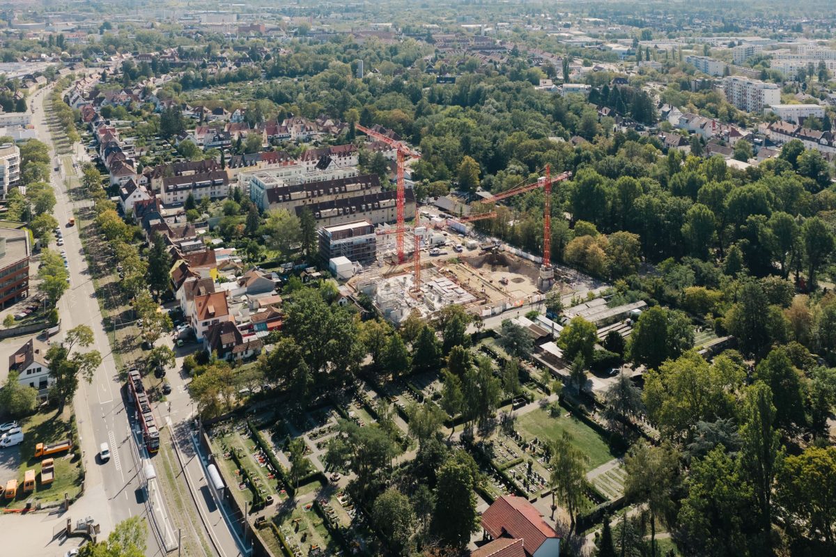 Luftaufnahme der Baustelle im Buschwiesenweg mit Kränen in einem Wohngebiet, umgeben von Bäumen, Straßen und Häusern an einem sonnigen Tag.