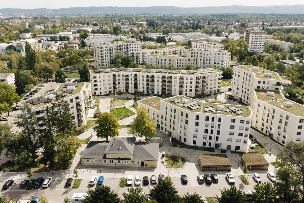 Luftaufnahme des August-Klingler-Areals mit begrünten Dächern. Im Vordergrund das denkmalgeschützte Vereinsheim sowie ein Stadtbild mit vielen Bäumen und fernen Hügeln im Hintergrund.