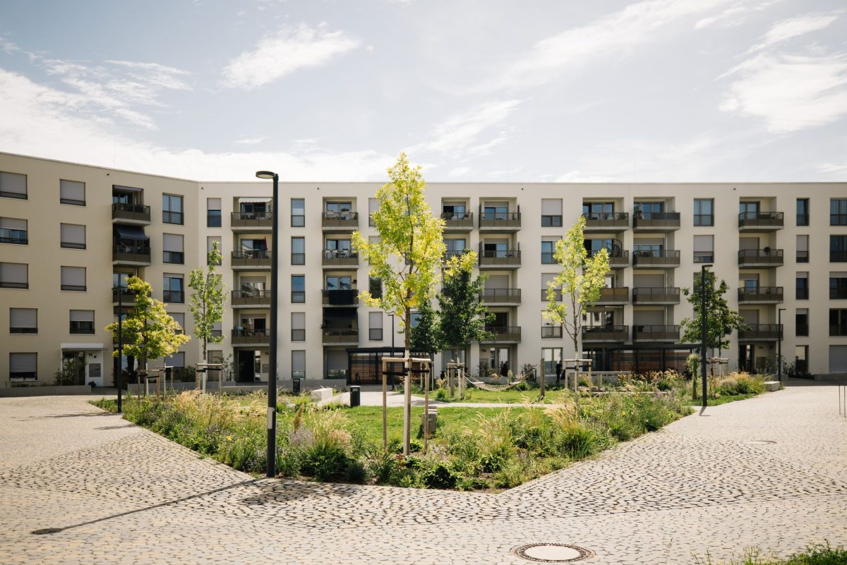 Ein modernes Mehrfamilienhaus mit Balkonen im August-Klingler-Areal, das einen zentralen Innenhof mit jungen Bäumen, Grünflächen und gepflasterten Wegen unter einem teilweise bewölkten Himmel umgibt.
