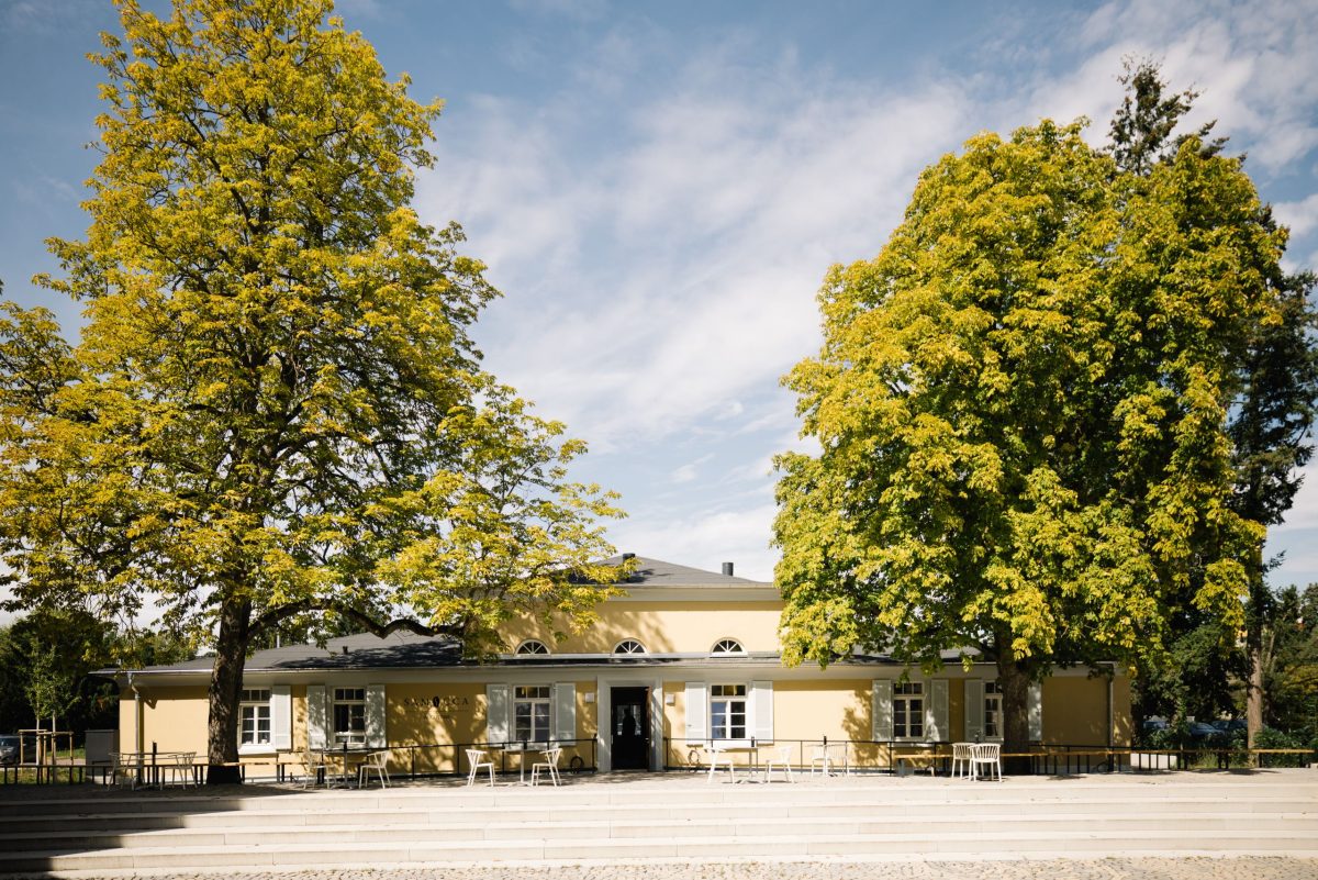Ansicht auf das denkmalgeschützte Vereinsheim im August-Klingler-Areal. Das Gebäude steht zwischen zwei großen, grünen Bäumen. Vor dem Gebäude sind Tische und Stühle im Freien aufgestellt.
